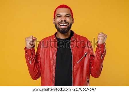 Young african fun man 20s with funky trendy pink hairdo in stylish red leather jacket black t-shirt do winner gesture clench fist celebrating isolated on yellow orange color background studio portrait Royalty-Free Stock Photo #2170619647