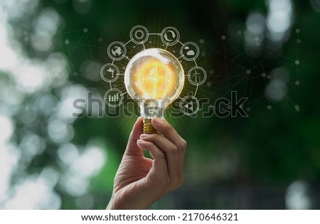 Hand holding light bulb with energy sources interface icons on green background, Technology and ecology sustainable development concept.