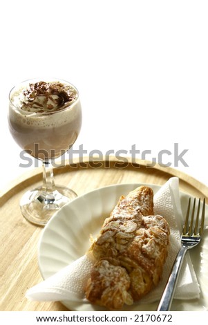A glass of  delicious hot chocolate/mocha coffee with swirl of cream and a light dusting of chocolate and a hot almond croissant on wooden mat.