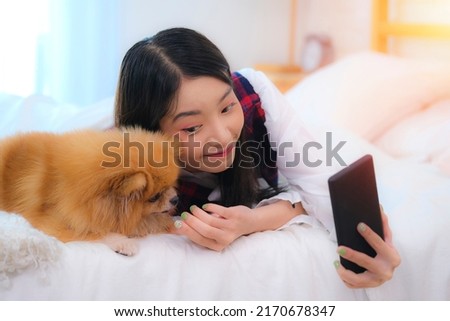 Beautiful young asian woman holding and hugging her dog in the room, Adorable asian woman smiling in blue and red seamless tartan plaid pattern shirt with dog and using smartphone to take a picture.