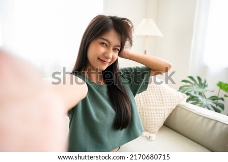 Pretty young asian female with big smile sitting at living room. She having fun taking light cheerful selfie on blurred background