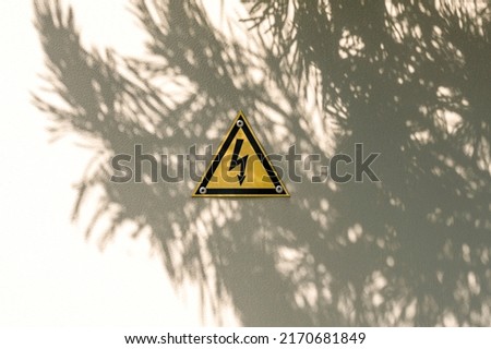 High voltage yellow triangle warning sign on gray metal doors with tree shadow. Danger electricity warning sign. Black lightning arrow symbol on yellow background in triangle.