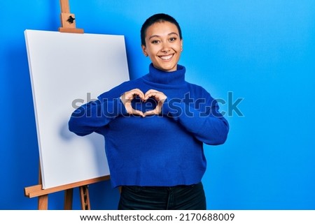 Beautiful hispanic woman with short hair standing by painter easel stand smiling in love showing heart symbol and shape with hands. romantic concept. 