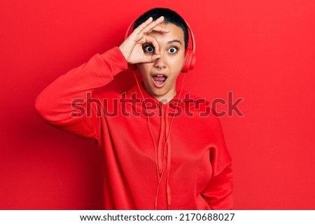 Beautiful hispanic woman with short hair listening to music using headphones doing ok gesture shocked with surprised face, eye looking through fingers. unbelieving expression. 