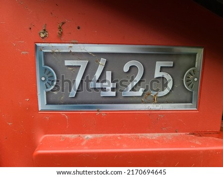 A closeup of the number 7425 against a red door. The house number is engraved into a metal plaque.