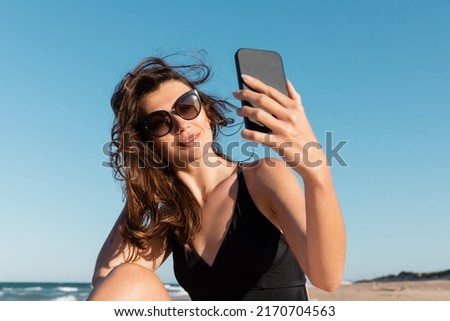 young woman in swimsuit and sunglasses taking selfie on smartphone on beach