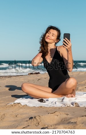 pleased young woman in swimsuit taking selfie on smartphone while sitting on beach