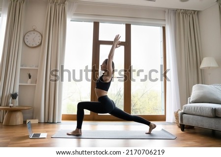Yoga instructor lead on-line class through video call application on laptop, standing in Warrior I pose practice Virabhadrasana, side full-length view. Sport work out at home, tech, quarantine concept Royalty-Free Stock Photo #2170706219