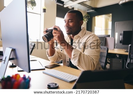 Young Black Professional Photographer Sitting At Desk Working On Computer Holding Camera Editing Pictures