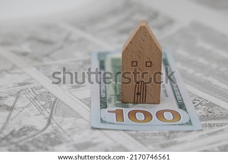 A wooden model house stands on a dollar bill against the background of a land plot scheme, real estate purchase