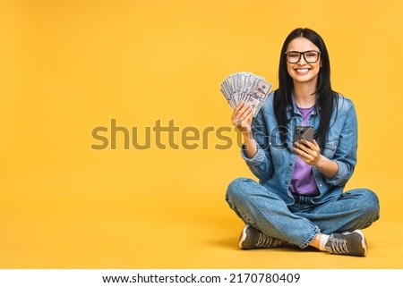 Happy winner! Portrait of a cheerful young woman holding money banknotes and celebrating victory isolated over yellow background. Sitting on the floor in lotus pose. Using mobile phone.