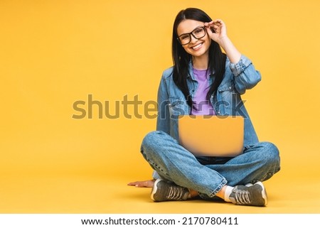 Business concept. Portrait of happy young woman in casual sitting on floor in lotus pose and holding laptop isolated over yellow background.