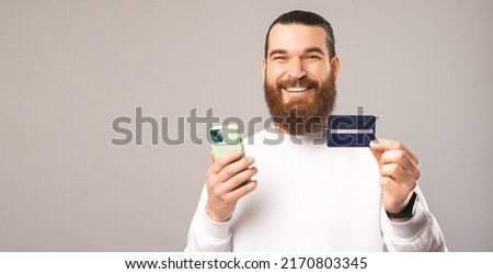 Panorama photo of a cheerful young bearded man holding a phone and a card. Studio shot over grey background.