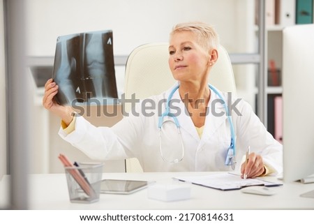 Pensive attractive mature radiologist with stethoscope on neck sitting at table and analyzing x-ray image on knee