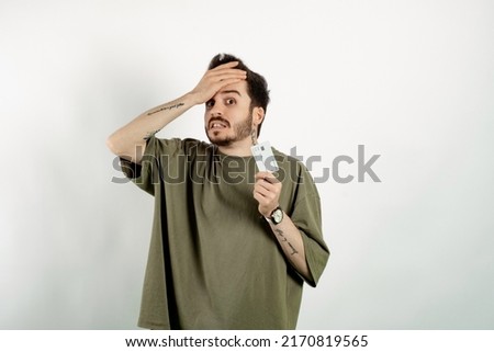 Caucasian young man wearing t-shirt posing isolated over white background showing credit card stressed with hand on head, shocked with shame and surprise face, angry and frustrated.