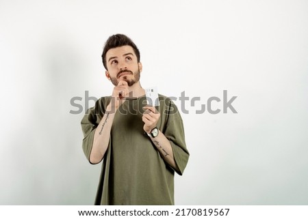Young man wearing khaki t-shirt posing isolated over white background holding credit or debit card serious face thinking about question, very confused ide