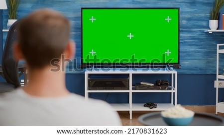 Over shoulder view of man watching movie on tv with green screen relaxing with bowl of popcorn sitting on couch. Back view of person relaxing on sofa in front of television mockup with chroma key