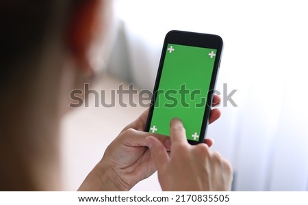 Chroma key mockup on smartphone in hand. Woman holds mobile phone iPhone and swipes photos or pictures left indoors of cozy home. Use green screen for copy space closeup