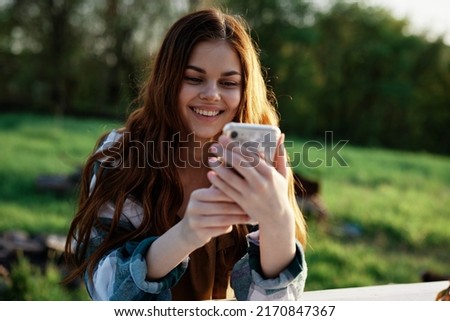 A beautiful woman relaxing and working on her phone sitting in nature in the park among the trees smiling and holding her smartphone in her hand lit by the bright sunset light Royalty-Free Stock Photo #2170847367
