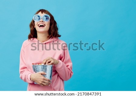 Movie concept. Happy cheerful cute redhead lady in pink hoodie sunglasses with popcorn posing isolated on blue studio background. Copy space Banner. Fashion Cinema. Entertainment offer