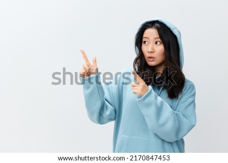 Cheerful happy cute Asian young female in blue hoodie sweatshirt point fingers up aside posing isolated on over white studio background. The best offer for ad. Fashion New Collection Ad concept