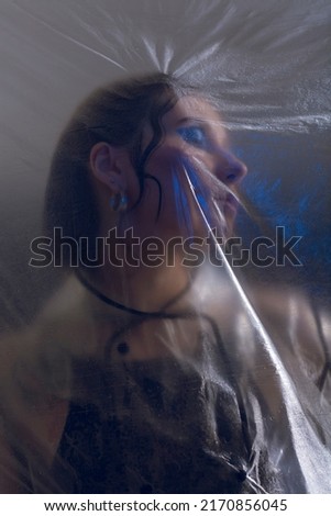 a photograph in blue is a portrait of a mysterious woman standing behind a plastic translucent film, pressing her face against it