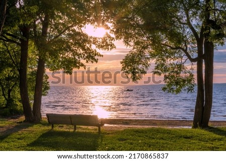 A pair of trees frames an empty park bench as a single boat floats on Lake Nipissing during a beautifully colourful sunset in North Bay, Ontario. Royalty-Free Stock Photo #2170865837