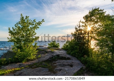 Sun beams poke their way through the lush green leaves of a tree on the shore of Lake Nippising during a beautifully colourful sunset in North Bay, Ontario. Royalty-Free Stock Photo #2170865849