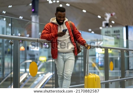 Joyful smiling young African American man using smartphone at arrivals lounge, happy black millennial guy standing with suitcase in terminal checking incoming messages after arrival to airport Royalty-Free Stock Photo #2170882587