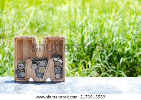 M-shaped wooden piggy bank on the back of green nature. Money saving concept. Copy space.