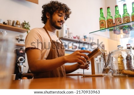 Young indian smiling handsome curly barista working with tablet standing behind the counter in a coffee shop Royalty-Free Stock Photo #2170933963