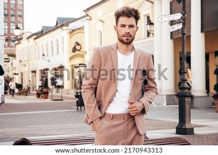 Handsome confident stylish hipster lambersexual model.Sexy modern man dressed in elegant beige suit. Fashion male posing in the street background in Europe city at sunset. In sunglasses Royalty-Free Stock Photo #2170943313