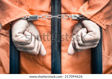 Handcuffed hands of a prisoner behind the bars of a prison with orange clothes - Crispy desaturated dramatic filtered look Royalty-Free Stock Photo #217094626