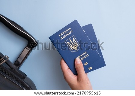 Passports of a citizen of Ukraine in a female hand on a blue background with suitcase, close-up. Emigration concept. Inscription in Ukrainian Ukraine Passport.