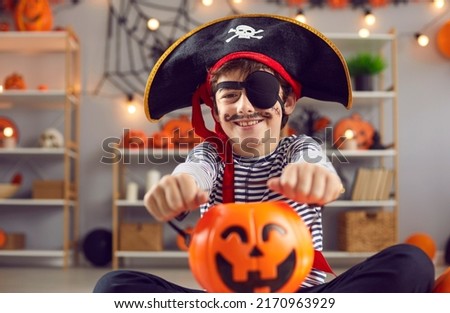 Indoor shot of happy child dressed up in kid's pirate costume holding orange Halloween pumpkin basket. Portrait of smiling little boy with fake painted mustache wearing black hat with skull and bones