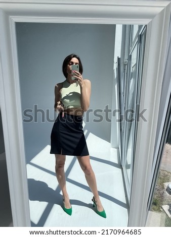 Fit tanned woman in classic short black skirt, light green crop top and heels, summer fashion stylish wear, take photo selfie on phone in mirror for social media, stories, vertical	