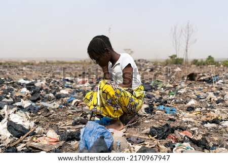 Tired little African girl who is desperately looking in the rubbish heap for objects that can be used; extreme poverty and hopelessness of the black continent Royalty-Free Stock Photo #2170967947