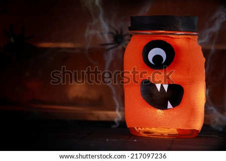 Orange jar decorated as a Halloween monster with a scary Cyclops face standing on rustic wooden shelves festooned in spiders and cobwebs on a shadowy background with copyspace