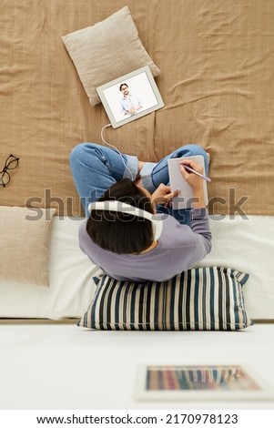 High angle view of young woman in headphones sitting on bed and making notes in notepad while watching webinar on tablet Royalty-Free Stock Photo #2170978123