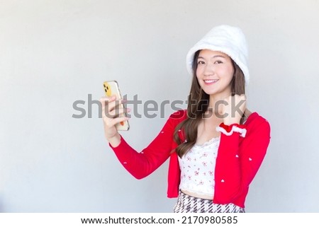 Beautiful young Asian woman who wears a red coat and white hat as a Santy girl holds a smartphone in her hand and another act while looking at the camera as glad and cheerful smiling.