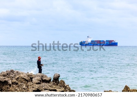 Fisherman standing on the rock and container ship sailing in sea  cloud sky background