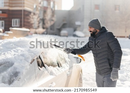 A young man cleans his car after a snowfall on a sunny, frosty day. Cleaning and clearing the car from snow on a winter day. Snowfall, and a severe snowstorm in winter. Royalty-Free Stock Photo #2170985549