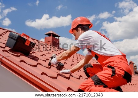 Young man worker master in red overall and helmet is fixing the metal tile roof.  Royalty-Free Stock Photo #2171012043