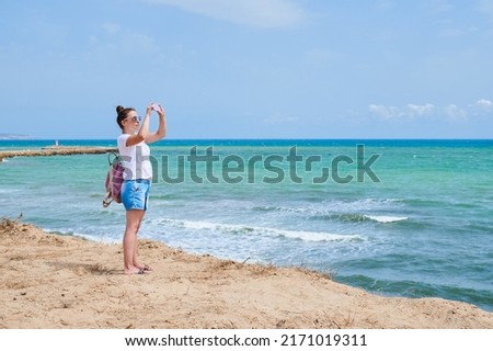 Young adult tourist woman with white shirt and jeans shorts takes pictures with her mobile from the top of a huge dune of a beautiful beach in Guardamar, Alicante, Spain, while enjoys summer vacation.