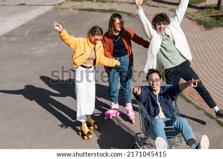 cheerful women riding roller skates near asian man showing rock signs in shopping cart