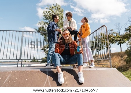 smiling woman with soda can sitting on skate ramp while multicultural friends talking on background