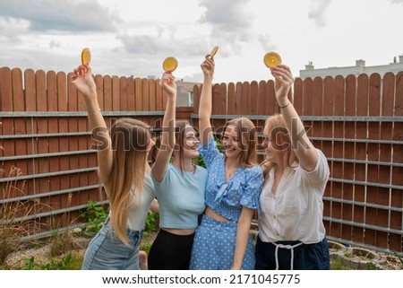 Laughing young girls standing together, raising hands with oranges and looking at each other, sky background. Having fun and enjoying outdoor recreation. Girls party, celebration, friendship.