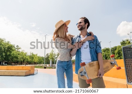Smiling woman in sun hat hugging boyfriend holding longboard in skate park