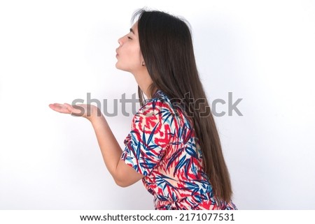 Profile side view view portrait of attractive young beautiful brunette woman wearing colourful dress over white wall sending air kiss