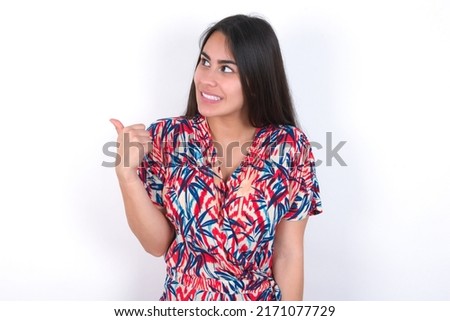 young beautiful brunette woman wearing colourful dress over white wall points away and gives advice demonstrates advertisement